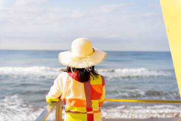 Unrecognizable woman from behind in colorful and striking dress and hat looks at the sea from the...