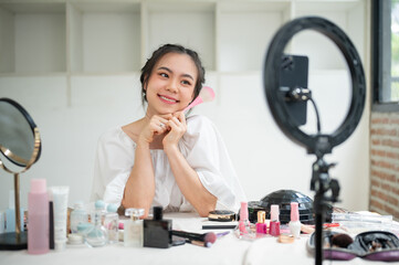 A cheerful Asian female beauty blogger recording her makeup tutorial video in her home studio.