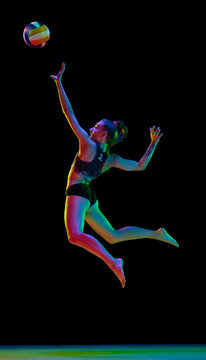 Dynamic full-length image of woman, professional female athlete playing volleyball against black studio background in neon light. Concept of professional sport, competition, health, hobby, action, ad