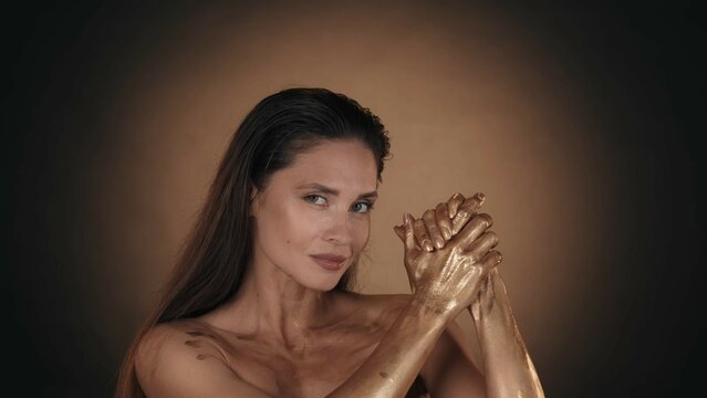 Attractive brunette model posing at the camera and applying golden liquid paint on her hands skin