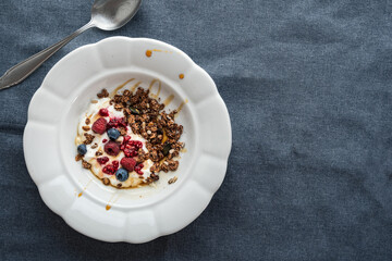 Ein Teller mit Granola, Joghurt und frischen Beeren auf einem blauen Tischtuch. Draufsicht.