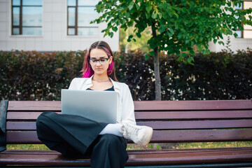 Obraz premium Charming long - haired female student using her laptop while sitting on the bench in the university campus. Beautiful young girl preparing for high school exams outdoors.