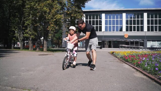 Father teaching child girl riding bike outdoors in a park. Involved hands-on father getting quality lime with child. 