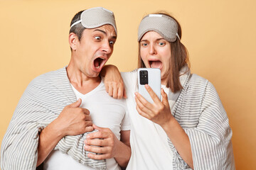Shocked couple man and woman wrapped in blanket isolated over beige background using mobile phone...
