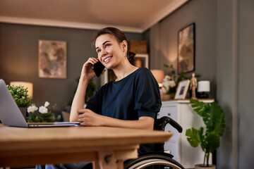 A woman in a wheelchair talking on a phone while working on a laptop at the home office.