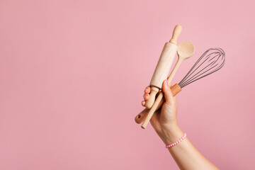Female hand holding kitchen utensils for food and bakery on pink background. whisk, rolling pin and...