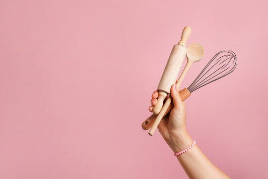 Female hand holding kitchen utensils for food and bakery on pink background. whisk, rolling pin and wooden spoon