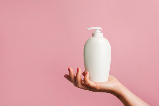 Woman holding white bottle of lotion, soap or shampoo in her hand, pink background with copy space
