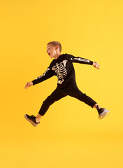 Obraz na płótnie Canvas Full-length image of little playful boy, child in black costume jumping against yellow studio background. Game. Concept of childhood, kids emotions, Halloween party, joy and fun, fashion, ad