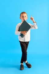 Ideas. Smart little boy, child in glasses, wearing white shirt and black pants standing with folders against blue studio background. Concept of childhood, education, school, kid emotions, ad