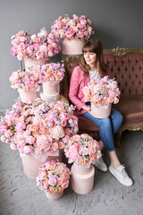 a happy young girl holds a box of flowers in her hands and inhales the fragrance of flowers. A beautiful woman is sitting on a sofa surrounded by flowers. A box with peonies and bush roses in a peach