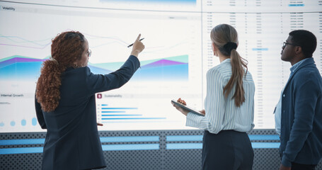 Diverse Team Of Data Scientists Analyzing Information And Graphs On Big Digital Screen In Modern Monitoring Office. Multiethnic Colleagues Discussing Financial Strategies, Risk Factors, and Deals.