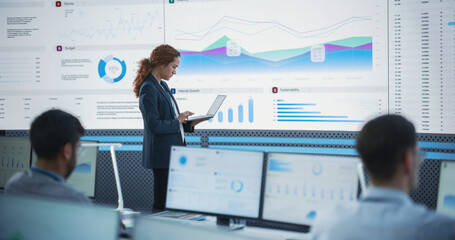 Female Project Manager Using Laptop And Analyzing Information Graphs On Big Digital Screen In Monitoring Office. Hispanic Woman Working For Big Data Company, Diverse Employees Behind Desktop Computers