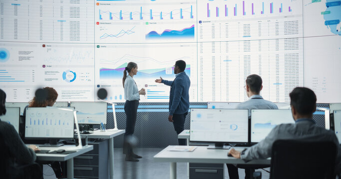 Black Male Financial Data Analysts And Caucasian Female Project Manager Having A Meeting In Monitoring Office With Analytics Feed On Big Digital Screen. Consulting Firm With Employees Behind Computers