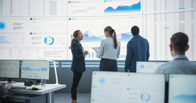 Diverse Team Of Financial Data Analysts Studying Information And Graphs On Big Digital Screen In Modern Monitoring Office. Multiethnic Employees Working Infront Of Desktop Computers In Consulting Firm - Powered by Adobe