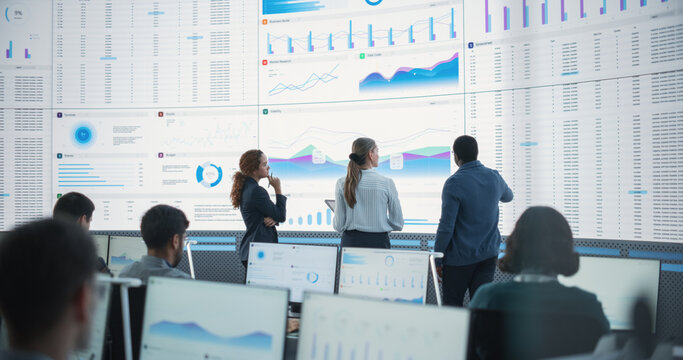 Diverse Team Of Data Scientists Having A Meeting And Discussing Reports In a Modern Monitoring Office With Analytics Feed On a Big Digital Screen. Big Data Company With Employees Behind Computers