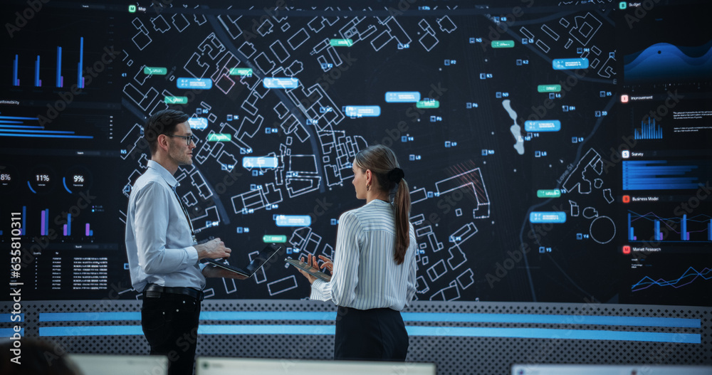 Poster Male And Female Data Analysts Discussing New Locations For Business In Front Of Big Digital Screen With City Map In Monitoring Office. Diverse Employees Working Behind Computers For Consulting Company