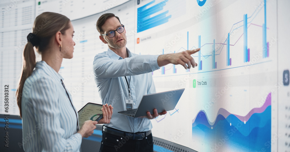 Poster caucasian male and female data scientists with laptop and tablet standing next to big digital screen