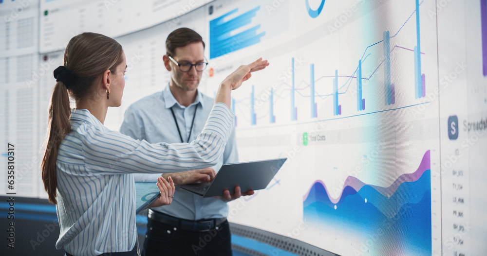 Poster caucasian male and female data scientists with laptop and tablet standing next to big digital screen