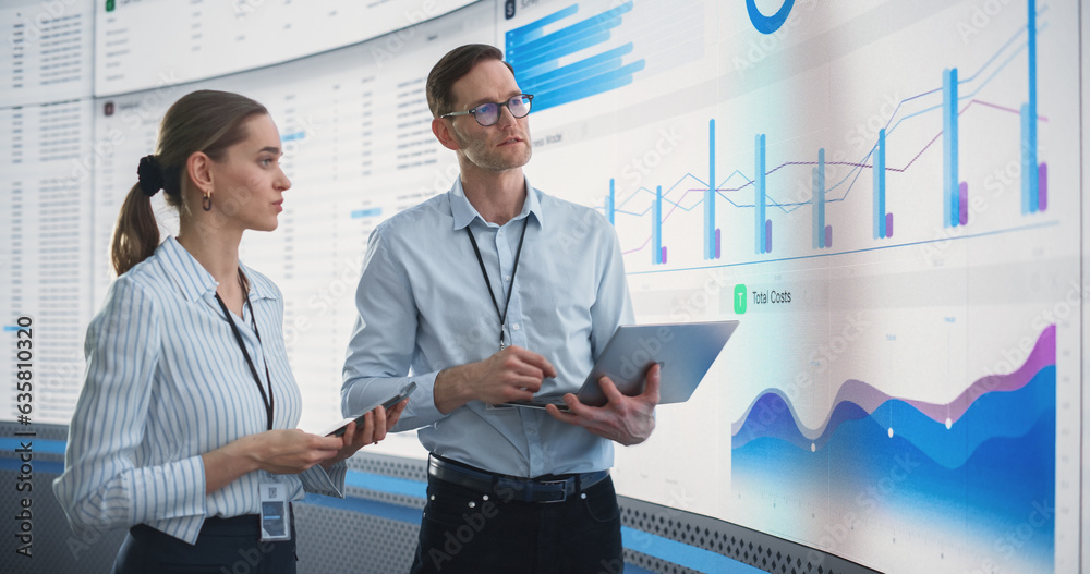 Poster Male And Female Data Analysts Discussing New Locations For Business In Front Of Big Digital Screen With City Map In Monitoring Office. Diverse Employees Working Behind Computers For Consulting Company