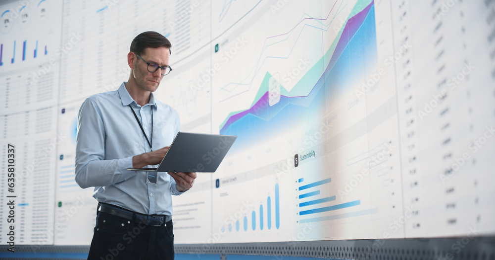 Poster In The Office: Male Monitoring Specialist Holding Laptop Computer And Standing Next To Big Digital Screen With Graphs And Charts. Caucasian Man Analyzing Data, Performance Records, And Filing Report.
