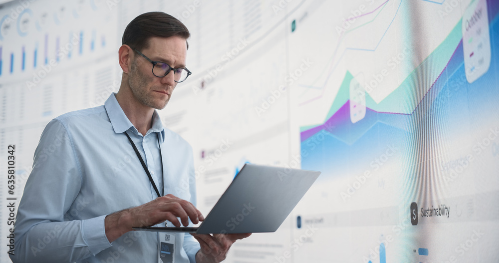 Poster in the office: male monitoring specialist holding laptop computer and standing next to big digital s
