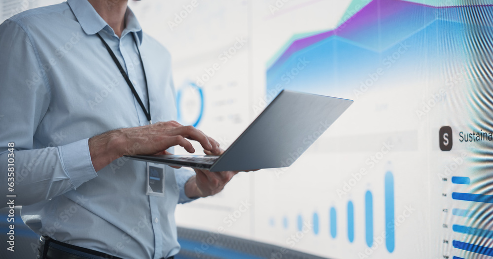 Fototapete Close Up Of Successful Caucasian Man Typing On Laptop Computer And Standing Next To Big Digital Wall With Data In The Office. Male Business Analyst Creating A Growth Strategy For Innovative Startup.