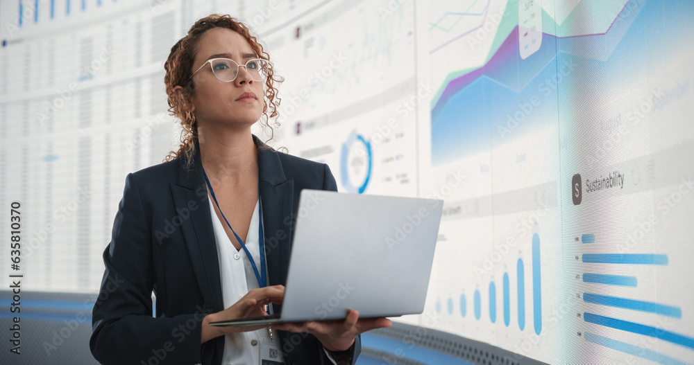 Poster In the Monitoring Office: Successful Hispanic Businesswoman Holding Laptop Computer And Analyzing Data On Big Digital Screen With Graphs and Charts. Female IT Entrepreneur Running Software Company.