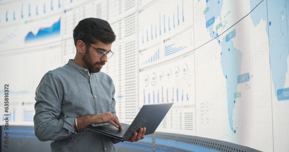 Poster indian male product distribution expert holding laptop computer, analyzing world map on big digital 