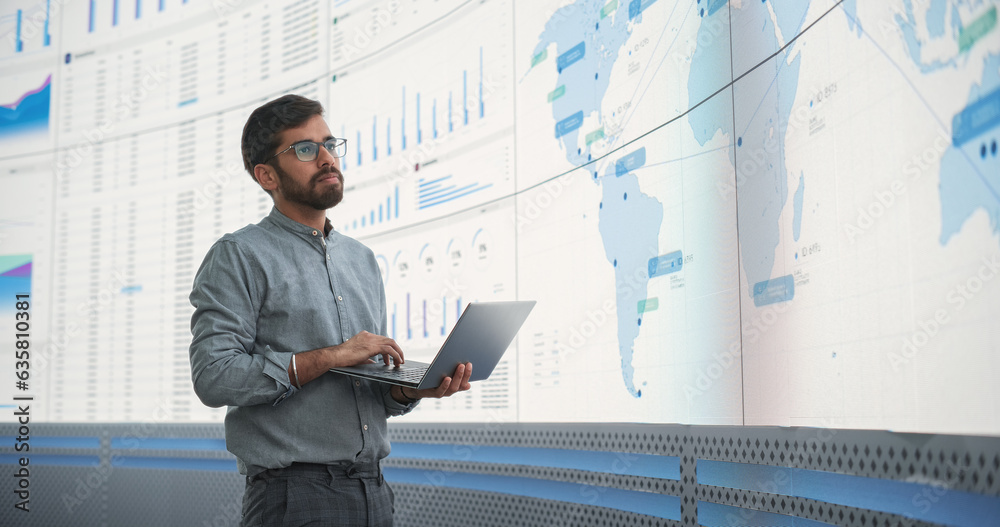 Poster indian male logistics expert holding laptop computer and analyzing world map on big digital screen i