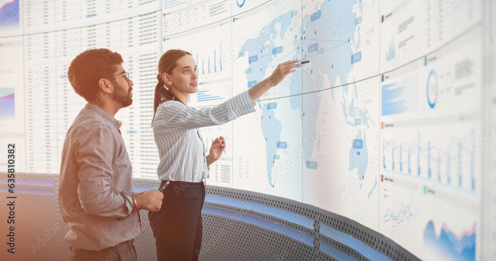 Leinwandbilder Indian Male Data Scientist And Caucasian Female Business Manager Talking In Front Of Big Digital Screen With Graphs, Charts And Map In Monitoring Office. Diverse Colleagues Discussing Objectives.