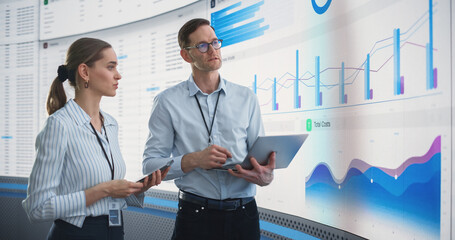 Male And Female Data Analysts Discussing New Locations For Business In Front Of Big Digital Screen With City Map In Monitoring Office. Diverse Employees Working Behind Computers For Consulting Company