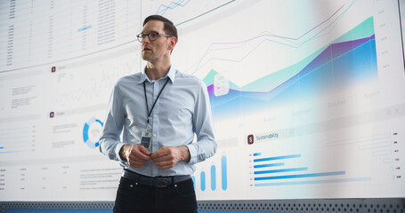 Male CEO Of Data Science Company Is Giving Presentation In Front Of Big Digital Screen With Graphs And Charts. Successful Caucasian Man Reviewing Business Accomplishments And Objectives In the Office.