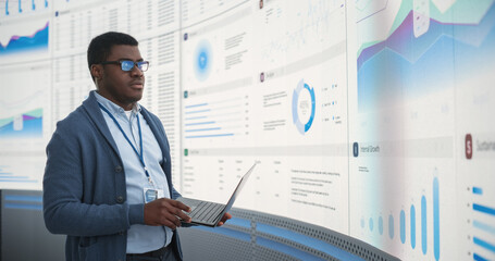Confident Black Businessman Holding Laptop And Reviewing Financial Reports On Big Digital Screen In...