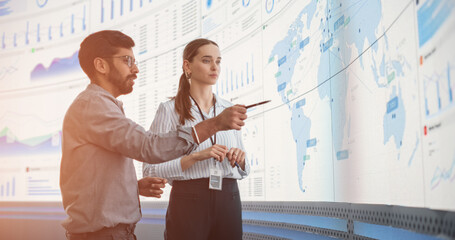 Indian Male Data Analyst And Caucasian Female Project Manager Talking In Front Of Big Digital Screen With Graphs, Charts And Map In Monitoring Office. Diverse Colleagues Discussing Profit Margins.