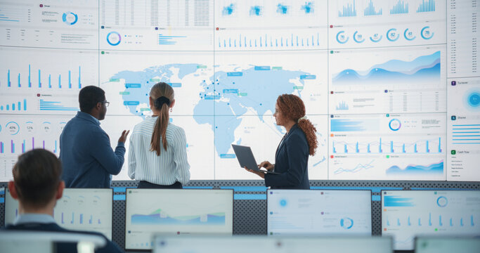 Diverse Team Of Specialists From Analytics Department Discussing Global Distribution In Front of Big Digital Screen With World Map In Monitoring Office. Multiethnic Employees Working Behind Computers.