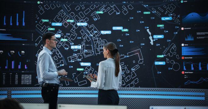 Male And Female Data Analysts Discussing New Locations For Business In Front Of Big Digital Screen With City Map In Monitoring Office. Diverse Employees Working Behind Computers For Consulting Company