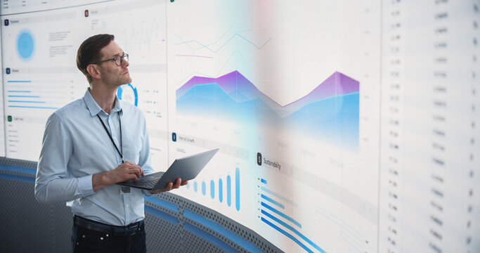 Caucasian Male Business Analyst Holding Laptop Computer And Standing Next To Big Digital Screen With Graphs And Charts. Successful Man Reviewing Financial Reports For Investment Management Company. - Powered by Adobe