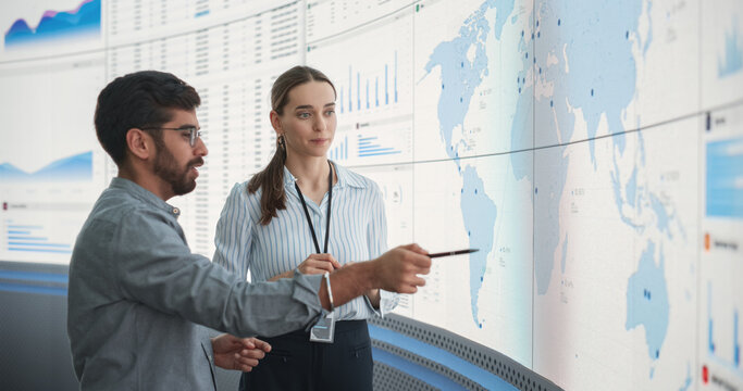 Caucasian Female Regional Manager Talking To Indian Male Distribution Expert In Front Of Big Digital Screen With Infographics And Map. Successful Man And Woman Discussing New Markets For Expansion.