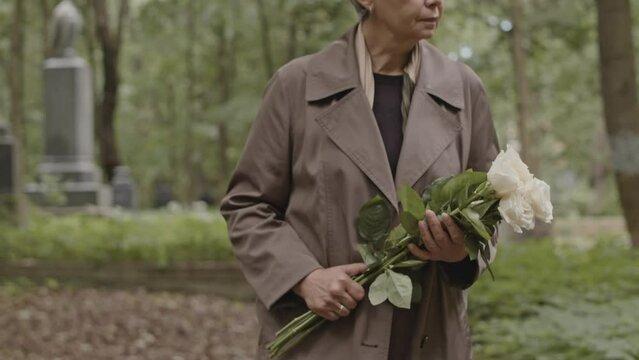 Cropped shot of unrecognizable woman with bouquet of white roses walking on graveyard alley at cemetery at daytime