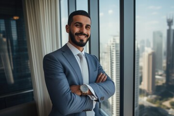 Fototapeta premium Young handsome Emirati business man in UAE traditional outfit standing on office with crossed arms. Arabic ambitious mature businessman.