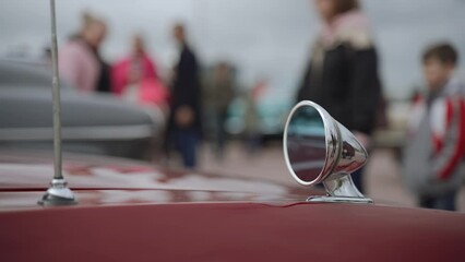 Side chrome bullet mirror and telescopic radio antenna on fender of red sports old coupe car. In parking lot, families in warm clothes walk near cars and examine them on cloudy day.