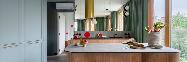 Modern composition of kitchen interior with wooden kitchen island, red barstools, colorful sculpture, green curtain, gold cooker hood, vase with rhubarb and personal accessories. Home decor. Template.