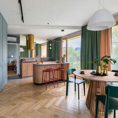 Interior design of harmonized dining room with kitchen, round table, green chairs, wooden kitchen island, gold cooker hood, brastools, vase with leaves and personal accessories. Home decor. Template.