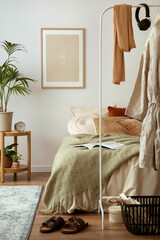 Warm and cozy composition of bedroom with mock up poster frame, green bedding, wooden bedside table, white hanger with clothes, black basket, gray carpet and personal accessories. Home decor. 