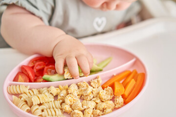 Obraz na płótnie Canvas Cute child eats healthy food pasta and vegetables steamed,. Portraits of a cute 10 months old baby girl. The baby sitting in a special high chair for babies. 