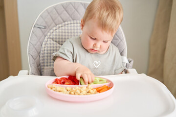 Cute child eats healthy food pasta and vegetables steamed,. Portraits of a cute 10 months old baby girl. The baby sitting in a special high chair for babies.  - 635824528