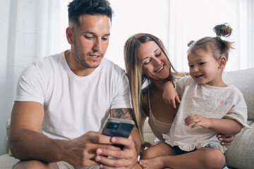 Father and mother using the mobile phone to put drawings or entertain their young daughter sitting on the sofa.