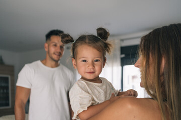 Fototapeta premium Portrait of a little girl with two lock of hair looking at the camera while being held by her mother and watched by the attentive gaze of her father.