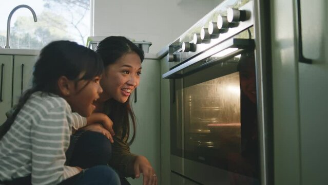 Asian mother daughter looking at cupcakes rise in the oven 